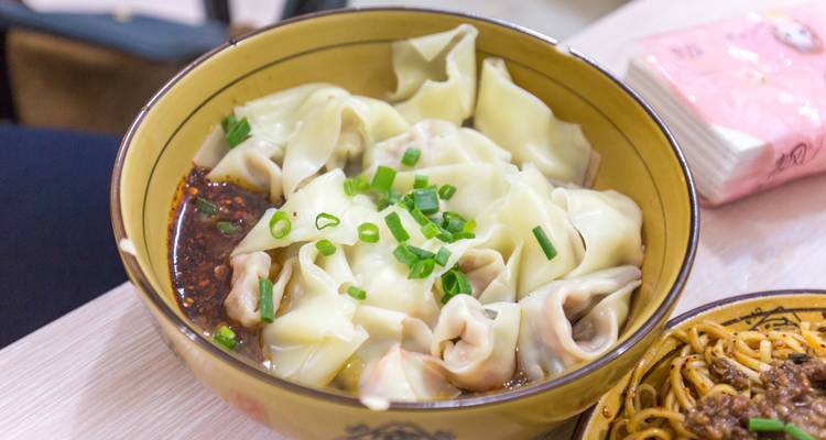 Bol de raviolis chinois dans un bouillon léger accompagné d'oignons verts hachés et d'une sauce épicée.
