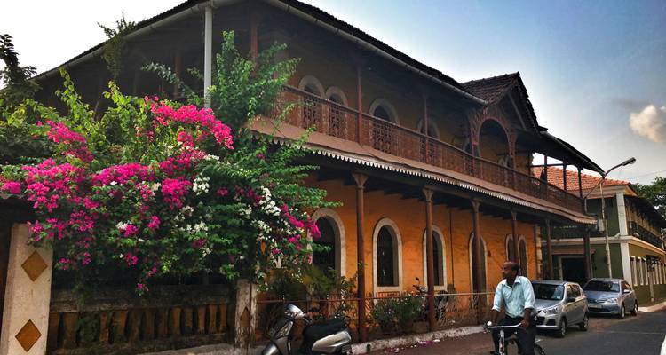 A scenic street scene in Goa, India, featuring an old colonial building with vibrant flowers and a person on a bicycle.