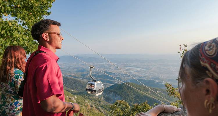 Los viajeros disfrutan de un mirador de teleférico de montaña con vista a un vasto valle bañado por la luz de la tarde.