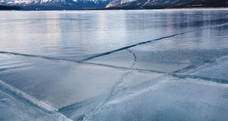 Lac gelé avec des fissures de glace géométriques reflétant des montagnes enneigées au loin en Alberta.