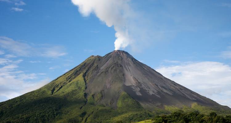 Dampf steigt aus dem Kegel des Arenal-Vulkans auf, der sich über grüne Regenwaldabhänge unter blauem Himmel erhebt.