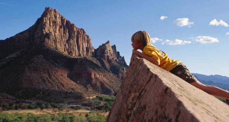 Un enfant escalade un rocher incliné pour admirer les imposantes falaises rouges du parc national de Zion.