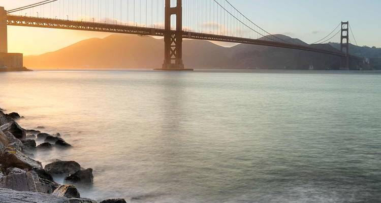 Puente Golden Gate silueteado al atardecer sobre las tranquilas aguas de la bahía con colinas distantes resplandecientes