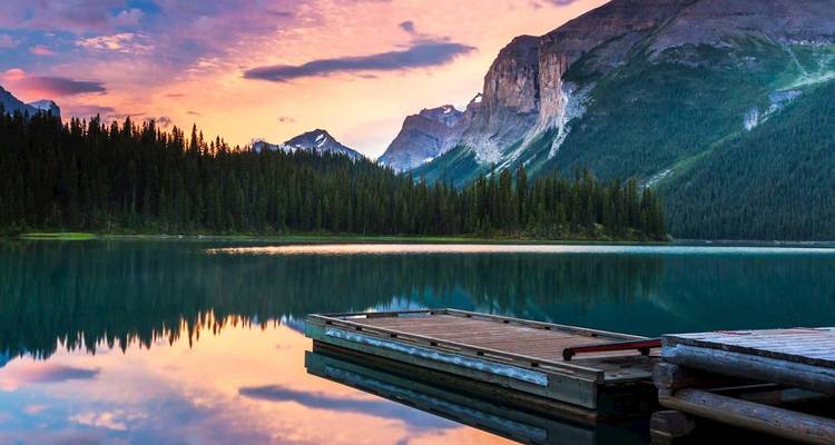 Lago alpino sereno al atardecer con un muelle de madera, orilla boscosa y dramático telón de fondo montañoso reflejado en aguas tranquilas.
