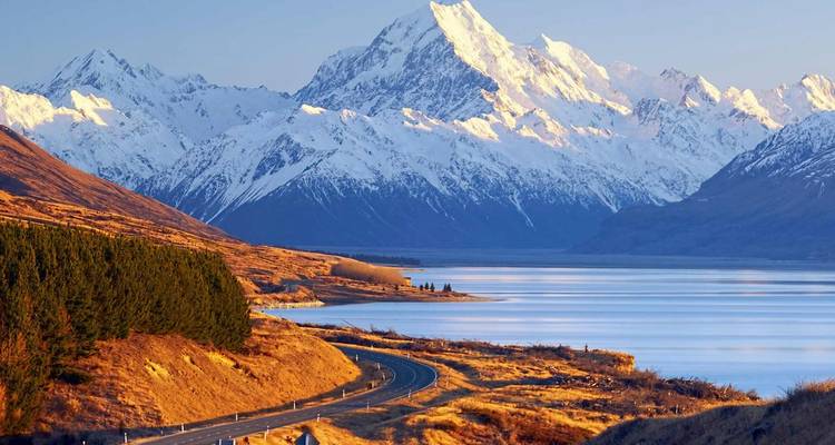 Le mont Cook recouvert de neige s'élève au-dessus d'un lac alpin tranquille et d'une route sinueuse baignée de lumière dorée.