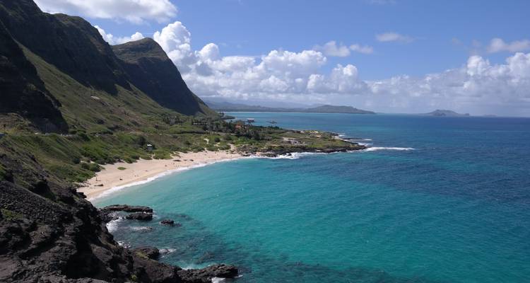 Een turquoise baai omzoomd door een zandstrand en steile groene kliffen onder een heldere blauwe hemel met verspreide wolken.