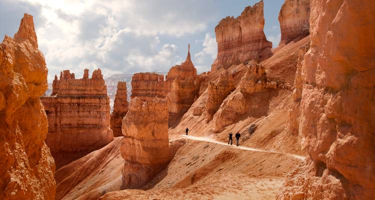 Los excursionistas caminan por un sendero polvoriento entre altas chimeneas de hadas naranjas en el Cañón Bryce bajo nubes suaves.