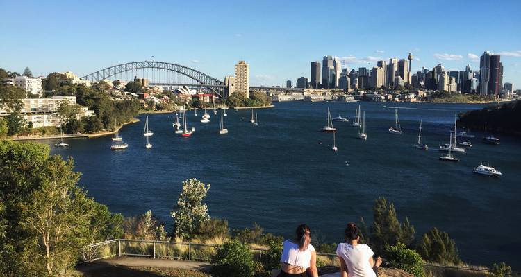 Zwei junge Frauen entspannen sich an einem Aussichtspunkt mit Blick auf den Hafen von Sydney mit der Harbour Bridge, der Skyline der Stadt und Segelbooten an einem sonnigen Nachmittag.