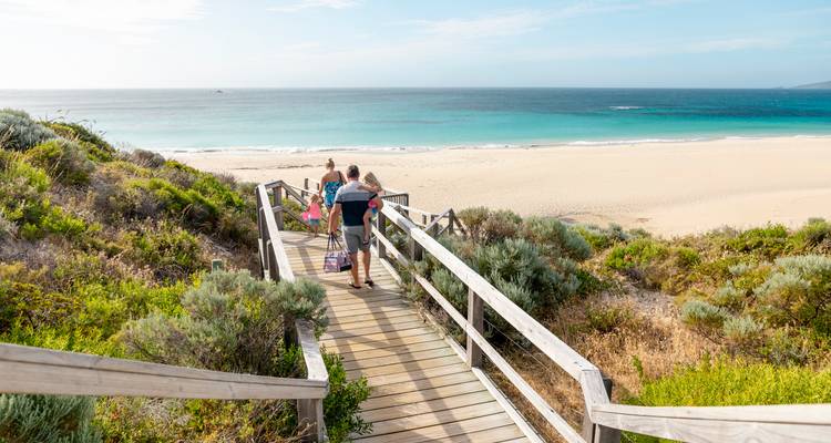 Familie trägt Strandausrüstung über einen hölzernen Steg hinunter zu einem breiten, leeren Sandstrand und türkisfarbenem Meer.