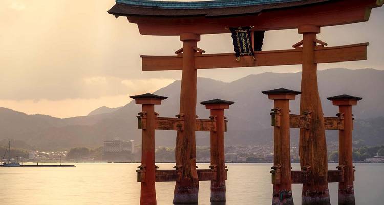 Iconic vermilion torii gate of Itsukushima Shrine rising from tranquil water against a backdrop of hazy mountains at golden hour.