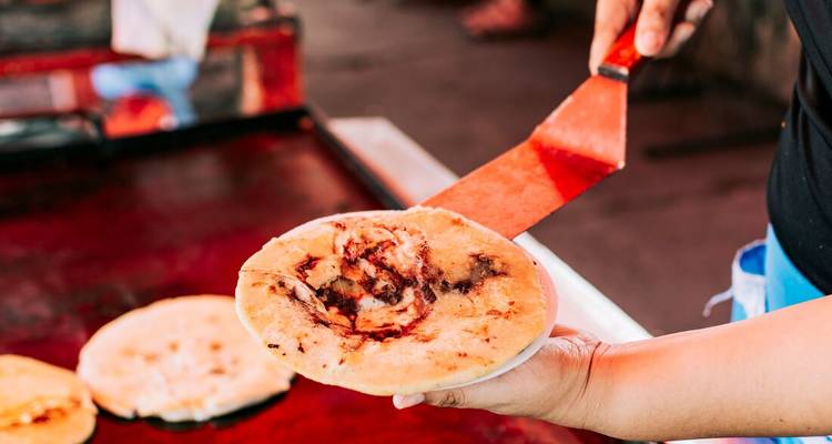 Straßenverkäufer wendet eine frisch gegrillte Pupusa mit einem roten Spatel über einer heißen Grillplatte