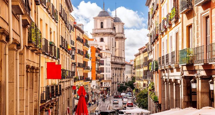 Calle estrecha y colorida en el distrito histórico de Madrid con balcones, torre, cafeterías y peatones.