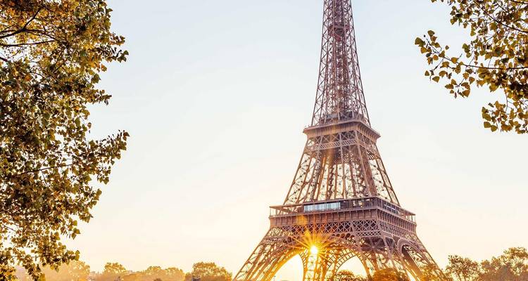 Eiffel Tower framed by autumn leaves with rising sun shining through its lattice.