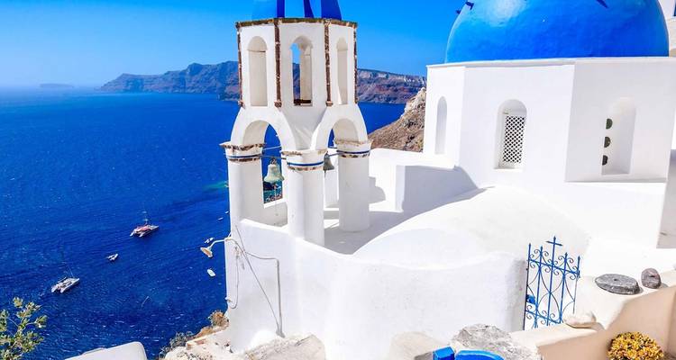 Iconische witte kerk met blauwe koepel en klokken met uitzicht op diepblauwe Egeïsche wateren op Santorini.