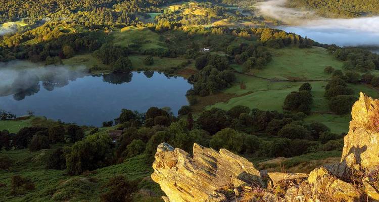 Un point de vue rocheux surélevé surplombe un petit lac rond entouré de champs verdoyants, de forêts et de la lumière brumeuse du matin