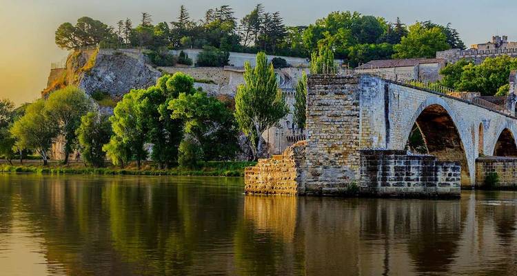 El histórico Pont d'Avignon y la fortaleza brillan con la cálida luz del atardecer reflejada en el río Ródano.