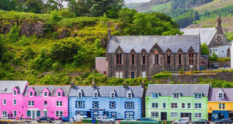 Rangée de cottages aux couleurs pastel au bord de l'eau et une église en pierre dans un décor de collines écossaises verdoyantes.