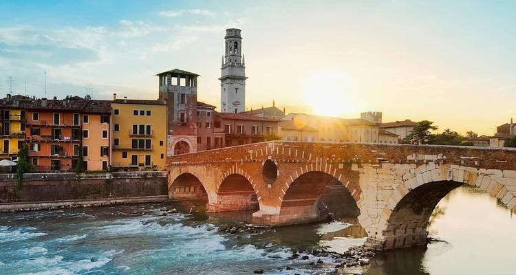 Verona’s stone Ponte Pietra bridge spans a rushing river at sunrise, historic towers and pastel houses behind