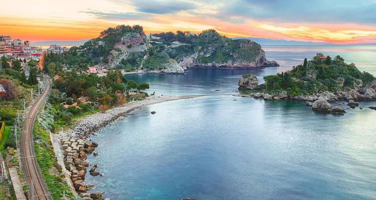 La baie turquoise, les îlots rocheux et la ville côtière de Taormina brillent au coucher du soleil avec les voies ferrées qui serpentent le long du rivage.
