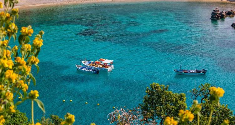 Kristalhelder turquoise baai met twee kleine bootjes omringd door bloeiende kustrotsen.