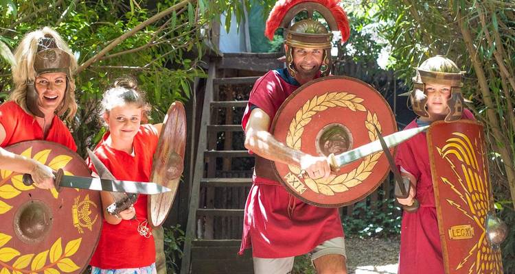 Familie, als römische Gladiatoren verkleidet, schwingt Holzschwerter in einer schattigen Umgebung im Freien.