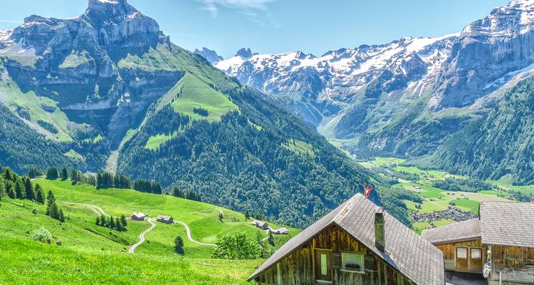 Grünes Alpental mit rustikalen Holzhütten unterhalb majestätischer schneebedeckter Schweizer Berge an einem hellen Tag