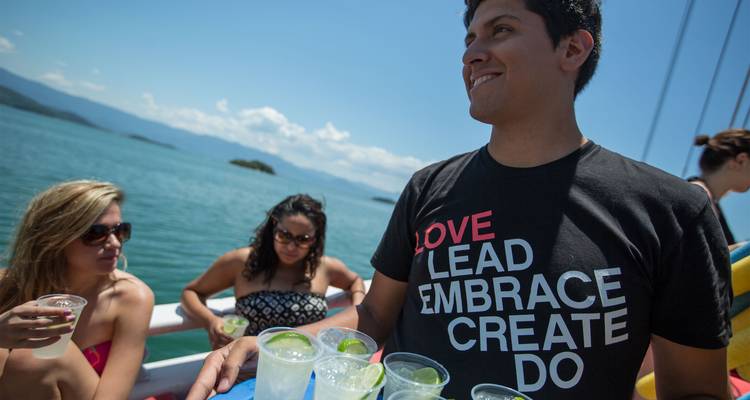 Jóvenes viajeros en un bote disfrutando de bebidas sobre aguas turquesas con islas boscosas a lo lejos.