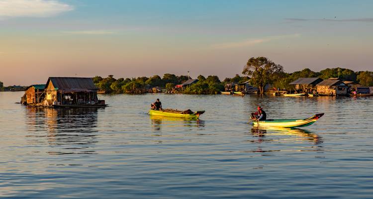 Des gens rament dans des bateaux sur une rivière calme, avec des maisons flottantes en arrière-plan au coucher du soleil.