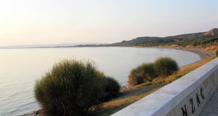 A tranquil coastline with shrubs and a stone wall with ANZAC engraved.