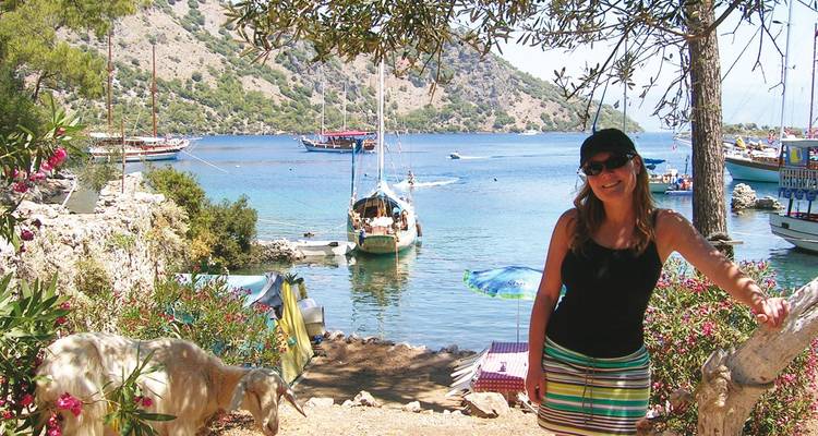 Smiling woman in summer outfit standing beside flowering shrubs overlooking a calm cove filled with boats; a goat grazes nearby.