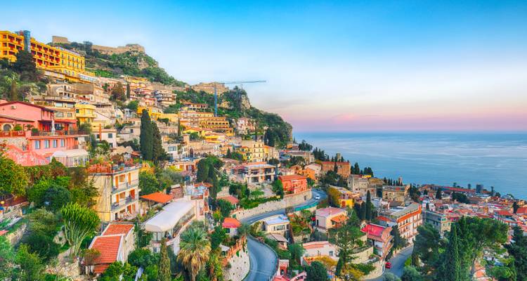Ville panoramique à flanc de colline de Taormina surplombant la mer calme à la lumière colorée du crépuscule.