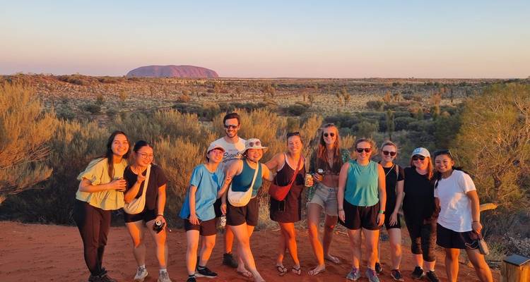 Un groupe de voyageurs se tient sur une dune de sable avec Uluru en arrière-plan à l'horizon à l'heure dorée.