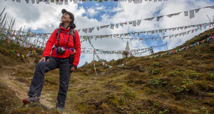Un senderista sonriente con una chaqueta roja se para entre banderas de oración ondeando en una ladera cubierta de pasto bajo nubes dispersas.