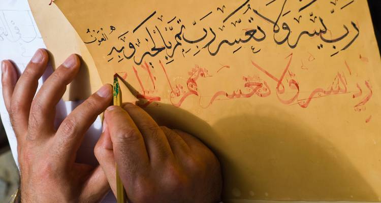 Hands practicing Arabic calligraphy with an ink pen on parchment.