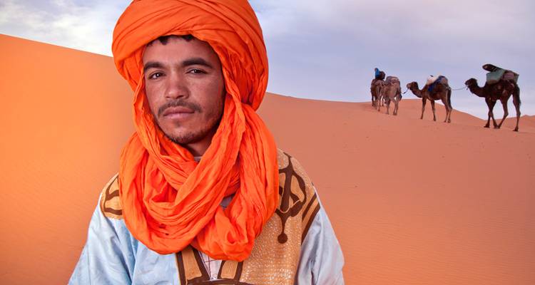 Young Berber man in bright turban stands before orange sand dunes and camel caravan.