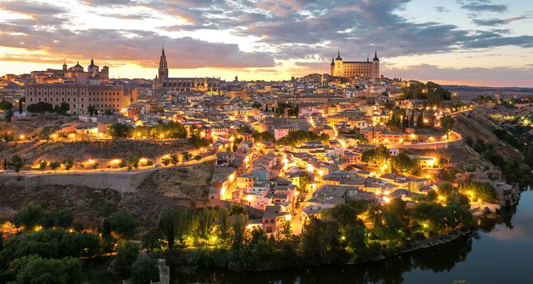 Vista panorámica del atardecer sobre la ciudad de Toledo iluminada en la colina con el río en primer plano.