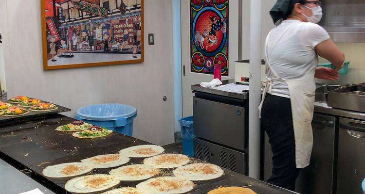 Un chef portant un masque prépare des crêpes de style street-food japonais dans une petite cuisine intérieure.