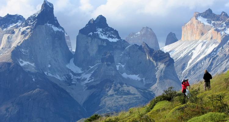 Zwei Wanderer stehen auf einem grünen Grat und blicken auf die dramatischen, schneebedeckten Granitürme der Torres del Paine