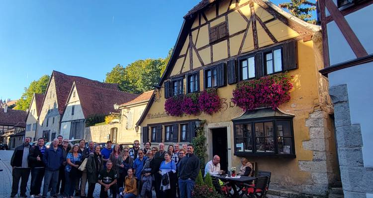 Grand groupe de touristes posant devant une maison à colombages ornée de fleurs roses dans une rue pavée.