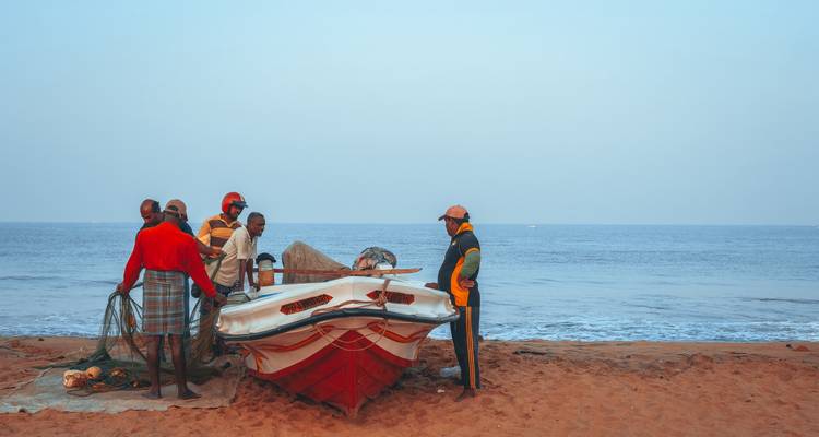Varios pescadores se reúnen alrededor de una pequeña barca roja y blanca en una playa de arena con un tranquilo mar azul de fondo.