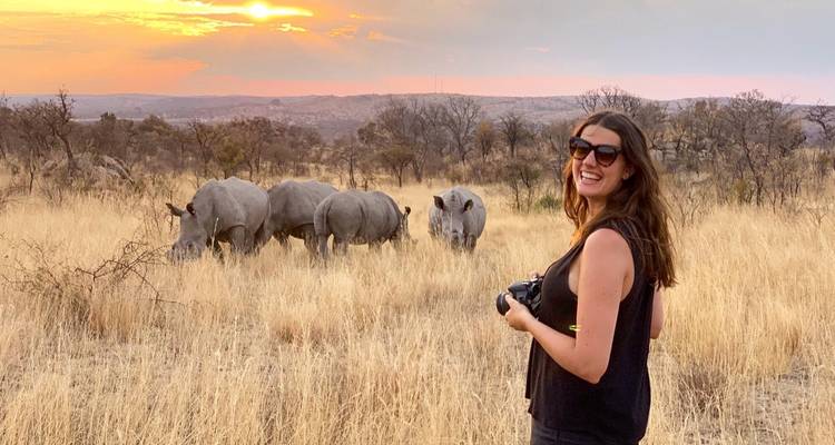 Femme avec un appareil photo près de rhinocéros qui broutent dans un paysage de savane au coucher du soleil.