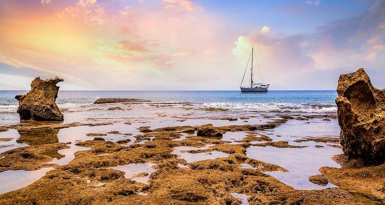 A sailboat on the horizon under a colorful sky over rocky shores.