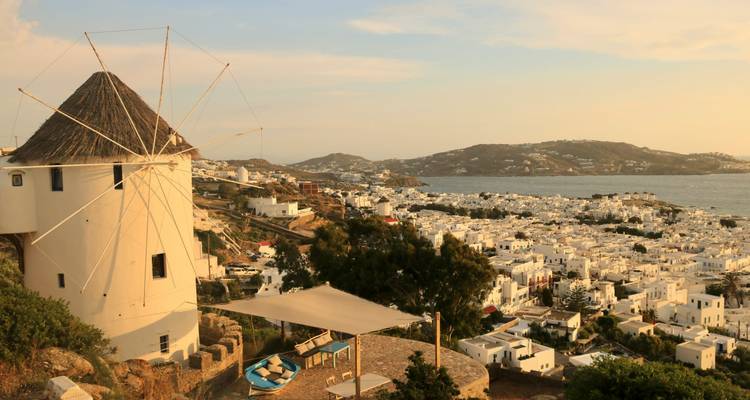 Een traditionele Mykonos windmolen kijkt uit over een uitgestrekt witgekalkt dorp en de glinsterende Egeïsche Zee bij zonsondergang.