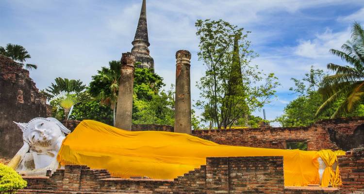 Riesige liegende Buddha-Statue, mit Safranstoff drapiert, inmitten alter Ziegelruinen mit einer aufragenden Chedi im Hintergrund.