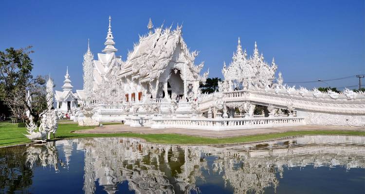 Glänzend weiße, kunstvoll verzierte Strukturen und Skulpturen des Wat Rong Khun, die sich in einem ruhigen Teich unter einem klaren blauen Himmel spiegeln.