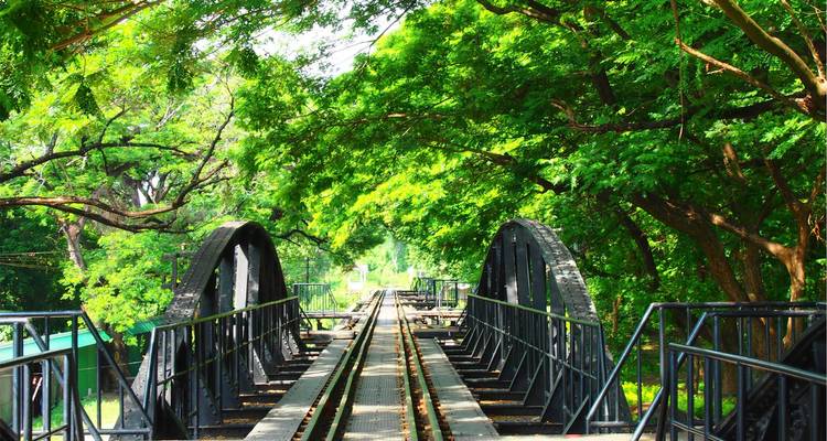 Stahlbogenbrücke über den Fluss Kwai, umrahmt von üppig grünem Baumbestand und Eisenbahnschienen, die sich in die Ferne erstrecken.