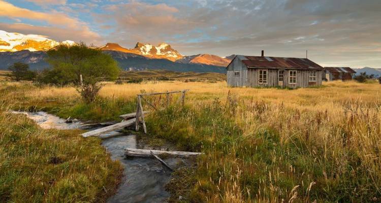 Eine von Witterung gezeichnete Hütte liegt in goldenen Graslandschaften, mit rauen Gipfeln, die im Abendlicht hinter ihr leuchten.