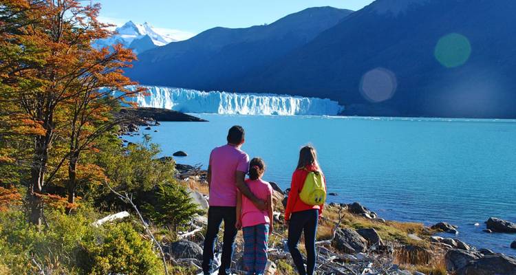 Eine dreiköpfige Familie steht neben einem türkisfarbenen See und blickt auf einen imposanten Gletscher und Berge.
