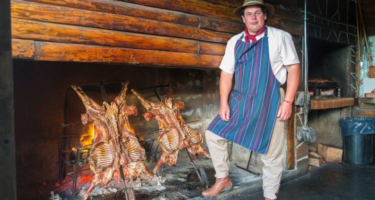 Ein Gaucho-Chef in einer Schürze steht neben ganzen Lämmern, die über einem offenen Feuer in einer rustikalen Küche braten.