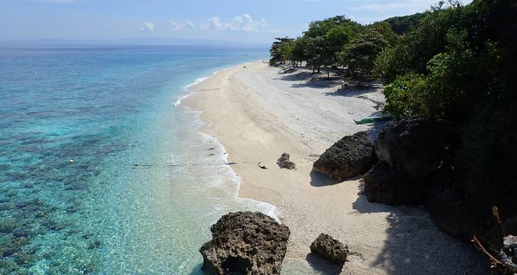 Crystal-clear turquoise water laps a narrow white coral beach fringed by green trees and rocky outcrops.
